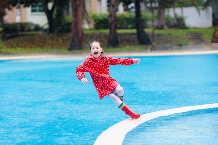 Kid jumping into swimming pool in rain. Child playing outdoors in tropical storm. Fun activities by summer rainy weather. Waterproof wear and footwear for kids. Children outdoors.の写真素材