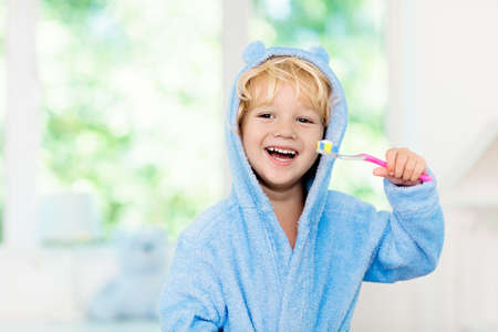 Child brushing teeth. Kids tooth brush and paste. Little baby boy in blue bath robe or towel brushing his teeth in white bathroom with window on sunny morning. Dental hygiene and heath for children.の写真素材