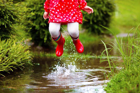 Kid playing in the rain in autumn park. Child jumping in muddy puddle on rainy fall day. Little girl in rain boots and red jacket outdoors in heavy shower. Kids waterproof footwear and coat.の写真素材