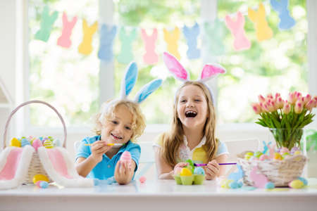 Kids dyeing Easter eggs. Children in bunny ears dye colorful egg for Easter hunt. Home decoration with flowers, basket and rabbit for spring holiday celebration. Little boy and girl decorate home.の写真素材