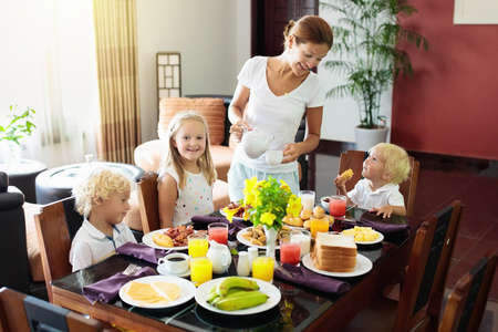 Healthy family breakfast at home. Mother and kids eating tropical fruit, toast bread, cheese and sausage. Children drink fresh pressed juice on sunny morning. Mom, boy, girl and baby eat breakfast.の写真素材