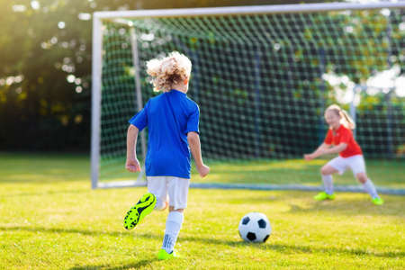 Kids play football on outdoor field. Children score a goal at soccer game. Girl and boy kicking ball. Running child in team jersey and cleats. School football club. Sports training for young player.の写真素材