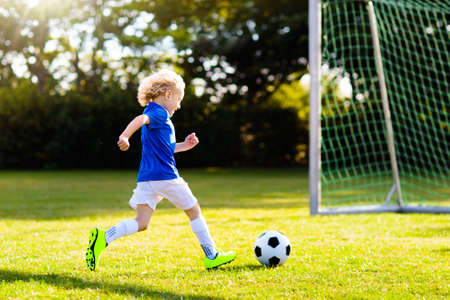 Kids play football on outdoor field. Children score a goal during soccer game. Little boy kicking ball. Running child in team jersey and cleats. School football club. Sports training for young player.の写真素材