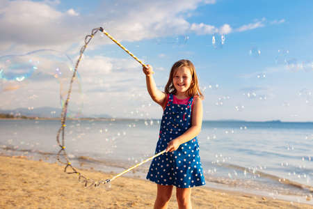 Kids blow bubbles at tropical beach. Child blowing soap bubble playing at sea. Family summer vacation with young kid. Outdoor beach activity for children. Water and sand toys for baby or toddler.の写真素材