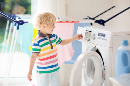 Child in a laundry room with washing machine or tumble dryer. Kid helping with family chores. Modern household devices and washing detergent in white sunny home. Clean washed clothes on drying rack.の写真素材