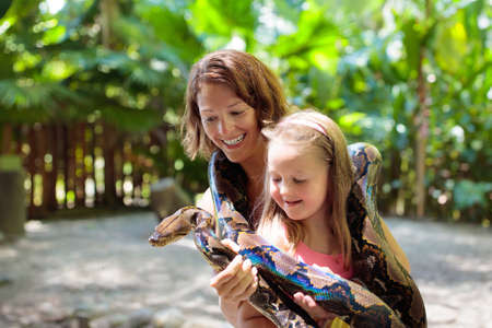 Kids holding python snake in tropical zoo. Family watching exotic reptile. Mother and child with...