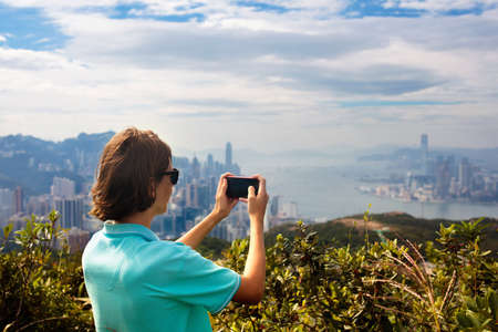 Young man hiking in Hong Kong mountains taking photo with smart phone camera of landscape with...