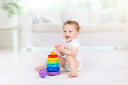 Baby playing with colorful toys. Educational toy stack for young child. Kids play at home. Little girl in white sunny nursery with pyramid and ball.の写真素材