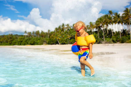 Child playing on tropical beach with palm trees. Little boy with inflatable ball and swim float at sea shore. Family summer vacation. Swimming aid for toddler. Kids play with water and sand toys.の写真素材