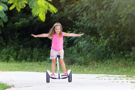 Child on hover board. Kids riding scooter in summer park. Balance board for children. Electric self balancing scooter on city street. Girl learning to ride hoverboard. Modern gadgets for school kid.の写真素材