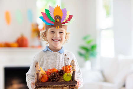Child celebrating Thanksgiving. Kid holding pumpkin in paper turkey hat. Autumn fun crafts and art. Little blond curly boy in decorated living room. Warm knitted wear. Fall season decoration.の写真素材