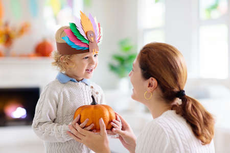 Child and mother celebrate Thanksgiving. Kid holding pumpkin in paper turkey hat. Autumn fun crafts and art. Mom and little boy in decorated living room. Warm knitted wear. Fall season decoration.の写真素材