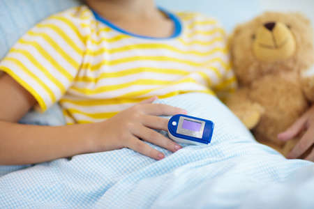 Sick little boy with pulse oximeter on his finger. Asthma treatment. Ill child lying in bed. Unwell kid with chamber inhaler, cough medicine. Flu season. Bedroom or hospital room for young patient.の写真素材