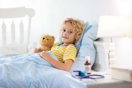 Sick little boy with asthma medicine. Ill child lying in bed. Unwell kid with chamber inhaler for cough treatment. Flu season. Bedroom or hospital room for young patient. Healthcare and medication.の写真素材
