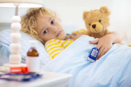 Sick little boy with pulse oximeter on his finger. Asthma treatment. Ill child lying in bed. Unwell kid with chamber inhaler, cough medicine. Flu season. Bedroom or hospital room for young patient.の写真素材