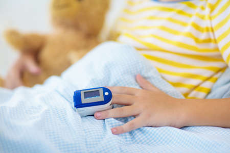 Sick little boy with pulse oximeter on his finger. Asthma treatment. Ill child lying in bed. Unwell kid with chamber inhaler, cough medicine. Flu season. Bedroom or hospital room for young patient.の写真素材