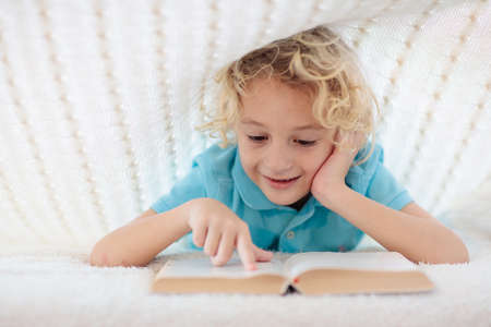 Child reading book in bed under knitted blanket. Kids cozy bedroom hygge style. Little boy doing...