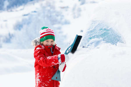 Child brushing snow off car after storm. Kid with winter brush and scraper clearing family car after overnight snow blizzard. Family Christmas vacation in the mountains. Kids shoveling snow.の写真素材