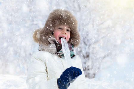 Child playing with snow in winter. Little boy in warm jacket and knitted hat catching snowflakes in...