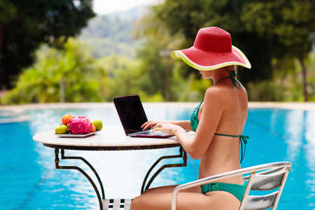 Woman with laptop at swimming pool. Remote work and global nomad concept. Young female freelancer in tropical resort. Conference call and freelance job. Girl in bikini with computer and mobile phone.の写真素材