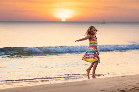 Child playing on ocean beach. Kid jumping in the waves at sunset. Sea vacation for family with kids. Little girl in beautiful dress running on tropical beach of exotic island during summer holiday.の写真素材