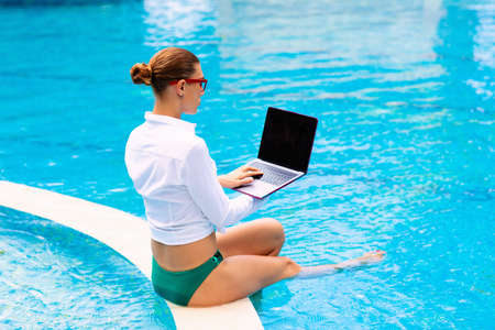 Woman with laptop at swimming pool. Remote work and global nomad concept. Young female freelancer in tropical resort. Conference call and freelance job. Girl in bikini with computer and mobile phone.の写真素材