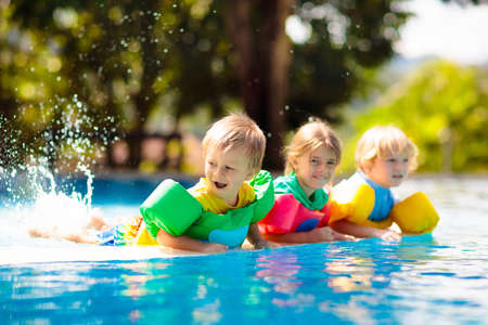 Kids play in outdoor swimming pool of tropical resort. Swim aid for young child. Baby learning to dive. Group of children playing in water. Colorful life jacket. Beach and summer fun.の写真素材