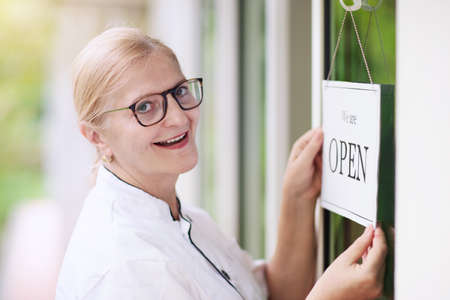 Cafe opening after lockdown. Open sign on front door of restaurant or grocery store. Waiter in apron greeting customer. Welcome message on bakery entrance. Back to work after quarantine.の写真素材