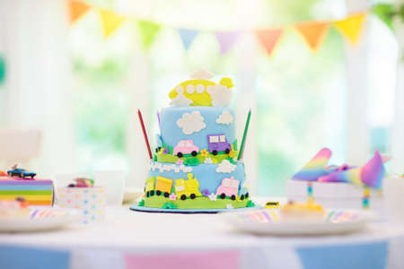 Kids birthday party. Boy cake with car and airplane. Child blowing out candles on colorful cake. Party decorations, rainbow flag banners, balloons. Vehicle theme celebration. Kid celebrating birthdayの写真素材