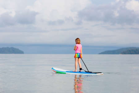 Child on stand up paddle board. Water fun and beach sport for kids. Healthy outdoor sports for summer vacation on tropical island. Holiday activity. Fit little girl training. Surfer exercising.の写真素材