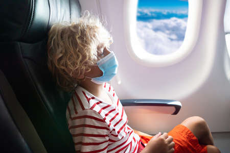 Child in airplane in face shield and mask. Flying with kids during virus outbreak. Vacation in coronavirus and flu pandemic. Safe travel with young child and baby. Kids fly airplane in surgical masks.の写真素材