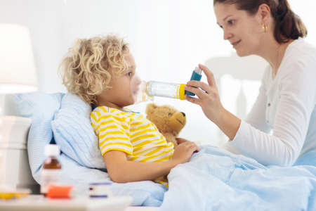 Sick little boy with asthma medicine. Mother with ill child lying in bed. Unwell kid with chamber inhaler for cough treatment. Flu season. Parent in bedroom or hospital room for young patient.の写真素材