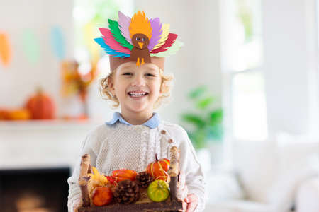 Child celebrating Thanksgiving. Kid holding pumpkin in paper turkey hat. Autumn fun crafts and art. Little blond curly boy in decorated living room. Warm knitted wear. Fall season decoration.の写真素材