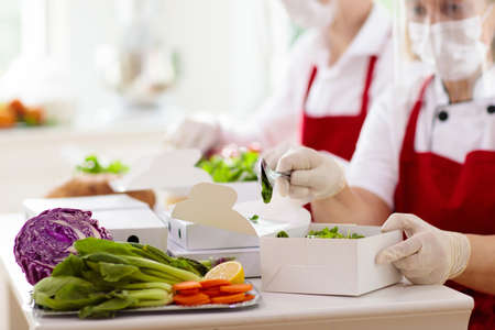 Food takeaway in restaurant in coronavirus outbreak. Cafe staff in face mask and shield cooking takeout order. Safety, covid-19 prevention. Chef with salad in cardboard container, eco friendly, vegan.の写真素材