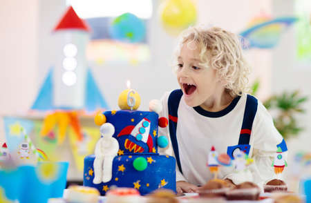 Kids space theme birthday party with cake and cupcakes. Rocket, solar system planet and spaceship decoration for child event. Little boy in astronaut costume blowing candles and opening presents.の写真素材