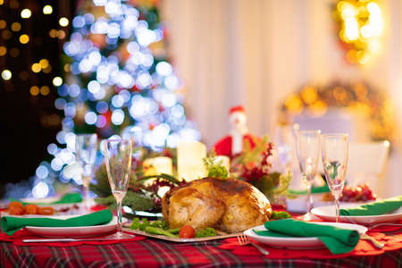 Christmas dinner at fireplace and decorated Xmas tree. Dish with roasted turkey, salad and baked potato served for festive family meal. Wine bottle with Santa hat. Open fire in stone oven.の写真素材