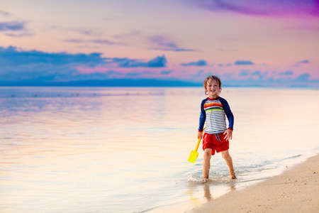 Child playing on ocean beach. Kid jumping in the waves at sunset. Sea vacation for family with kids. Little boy on tropical beach of exotic island on summer holiday. Sunset view. Sand and water fun.の写真素材