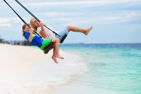 Child on beach swing. Kid swinging on tropical island. Travel with young children. Summer family exotic vacation. Kids play on sea shore. Little girl and boy playing in luxury ocean resort.の写真素材
