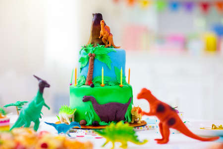 Kids birthday party. Dinosaur theme cake. Little girl blowing candles and opening gifts. Children event. Decoration for dinosaurs themed celebration.の写真素材