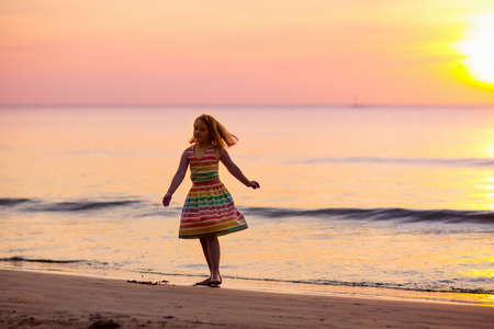 Child playing on ocean beach. Kid jumping in the waves at sunset. Sea vacation for family with kids. Little girl in beautiful dress running on tropical beach of exotic island during summer holiday.の写真素材