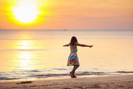 Child playing on ocean beach. Kid jumping in the waves at sunset. Sea vacation for family with kids. Little girl in beautiful dress running on tropical beach of exotic island during summer holiday.の写真素材