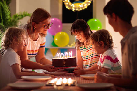 Kids birthday party. Child blowing candles on cake and opening presents. Pastel rainbow theme celebration. Family celebrating at home. Boy and girl open gifts, eat cakes. Sweets for children.の写真素材