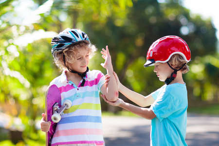 Kids with skate board. Children ride skateboard. Sports safety, fall and injury. Little boy hurt his knee and elbow after outdoor sport and exercise activity. Sibling or friend support and help.の写真素材