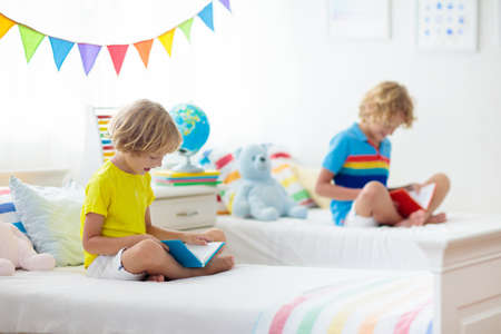 Child reading book in bed. Bedroom for two kids. Little boys read books on white bed with colorful bedding. Children nursery. Kid boy siblings play indoors. Nap and sleep time. Student doing homework.の写真素材