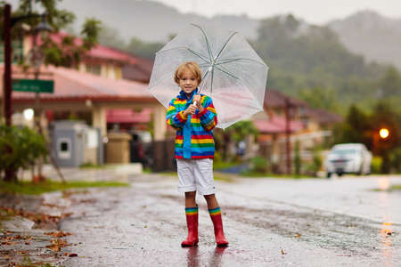 Child playing in autumn rain. Kid with umbrella. Little boy running on city street in fall season. Outdoor fun for kids by any weather. Rain waterproof wear, boots and jacket for children.の写真素材