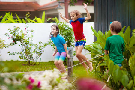 Kids playing with water hose in summer garden. Child water sprinkler fun on hot sunny day. Children jump and run in the backyard. Brother and sister play outside. Healthy outdoor activity for child.の写真素材