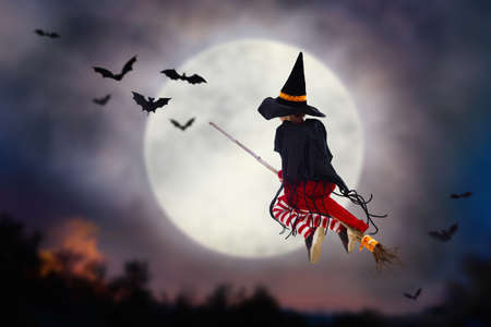 Little witch flying broom on Halloween night. Huge full moon and bats in the background. Kids trick or treat costume. Children have fun. Spooky and scary celebration. Kid dressed as evil ghost.の写真素材