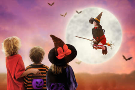 Little witch flying broom on Halloween night. Kids watch huge full moon and bats in dark window. Child trick or treat costume. Children have fun dressed up. Spooky and scary celebration. Kid at home.の写真素材