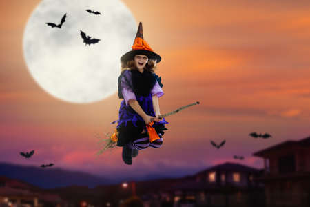 Little witch flying broom on Halloween night. Huge full moon and bats in the background. Kids trick or treat costume. Children have fun. Spooky and scary celebration. Kid dressed as evil ghost.の写真素材