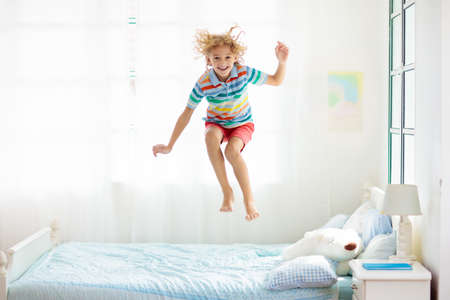 Child jumping on bed in white bedroom with big window. Kids play at home. Fun jump on parents bed. Room and interior design. Boy at home. Sunny morning, weekend family relaxing time.の写真素材